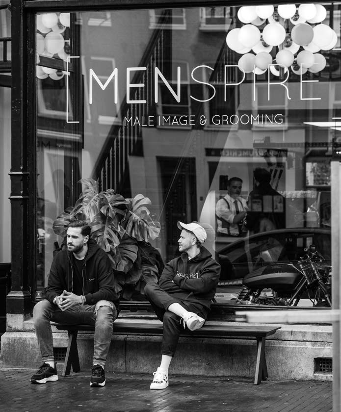 Samuel Palmer (à gauche) et Josh Lamonaca (à droite) assis sur un banc devant la vitrine d’un établissement MENSPIRE ; on lit sur la vitre « MENSPIRE – Male Image & Grooming ». Photo en noir et blanc.