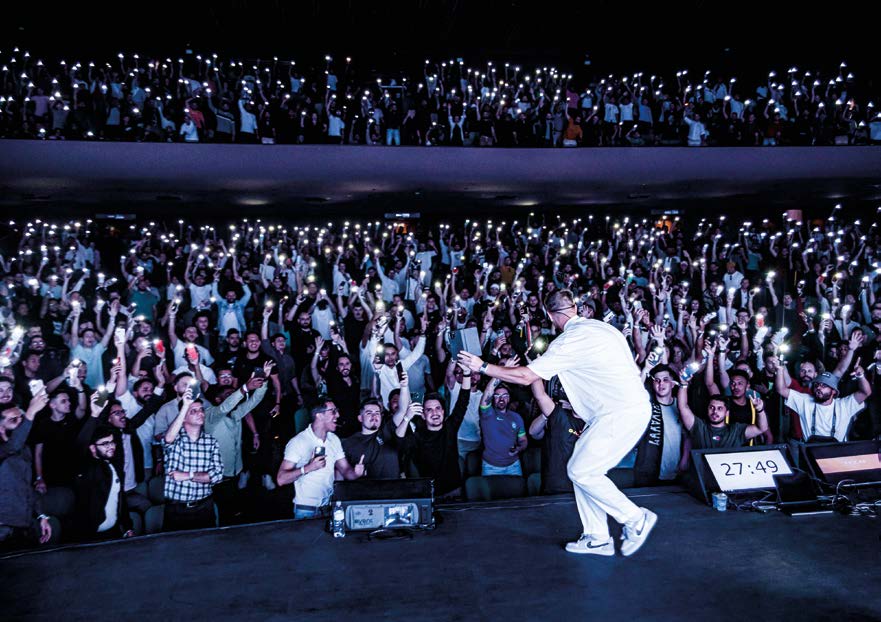 Un homme vêtu de blanc sur une scène, dos à l’objectif, face à une salle comble ; le public brandit les lampes des téléphones.