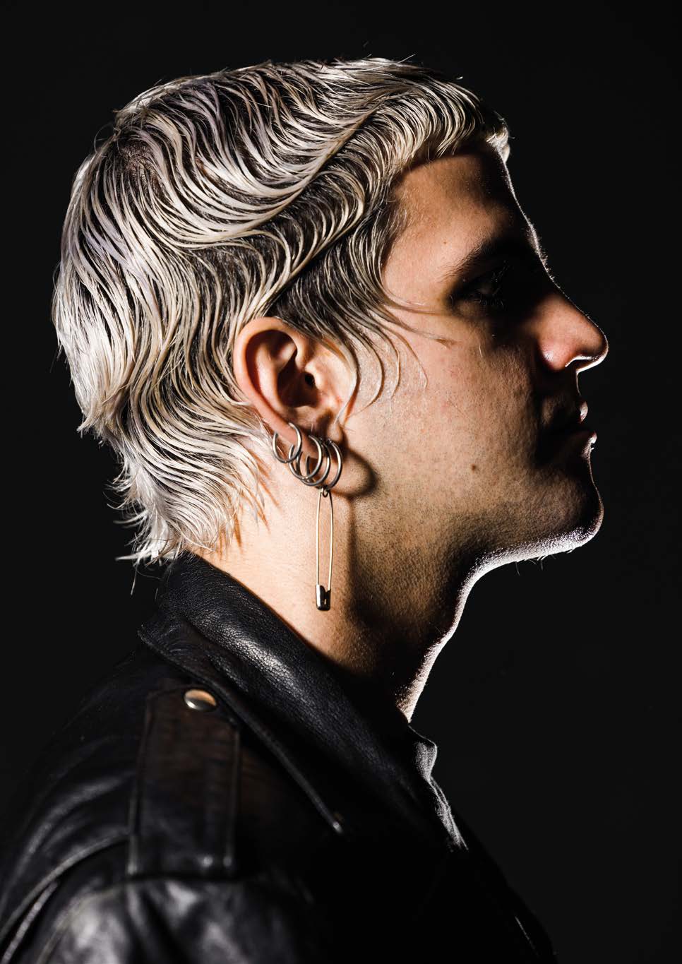 Modèle de profil aux cheveux décolorés et ondulés, peignés en arrière effet mouillé ; anneaux d’oreille avec une épingle de sûreté, blouson de cuir noir, fond noir.