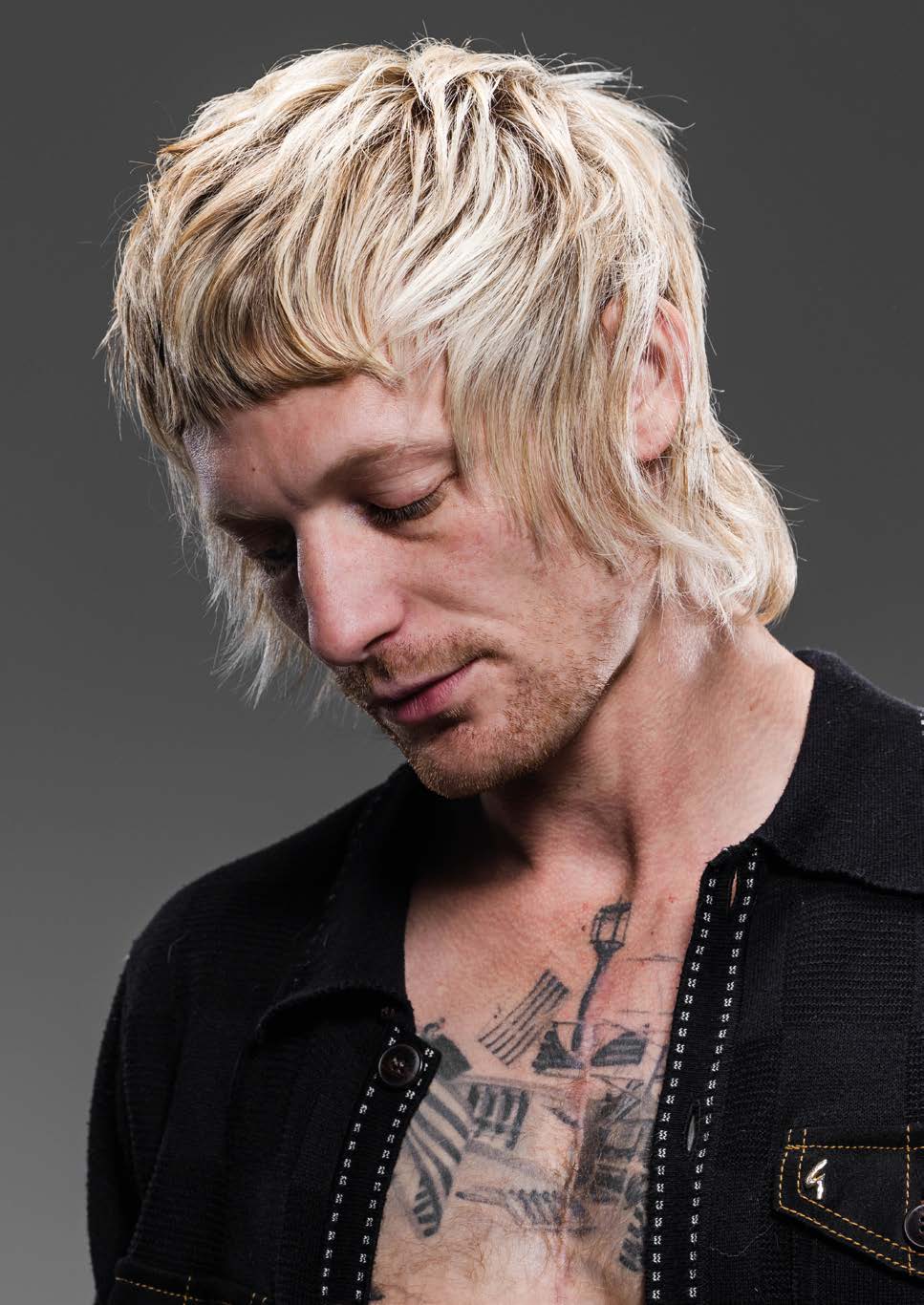 Modèle à la coupe blonde dégradée, frange courte et longueurs dans la nuque, le regard baissé ; gilet noir ouvert, tatouage sur le torse, fond gris.
