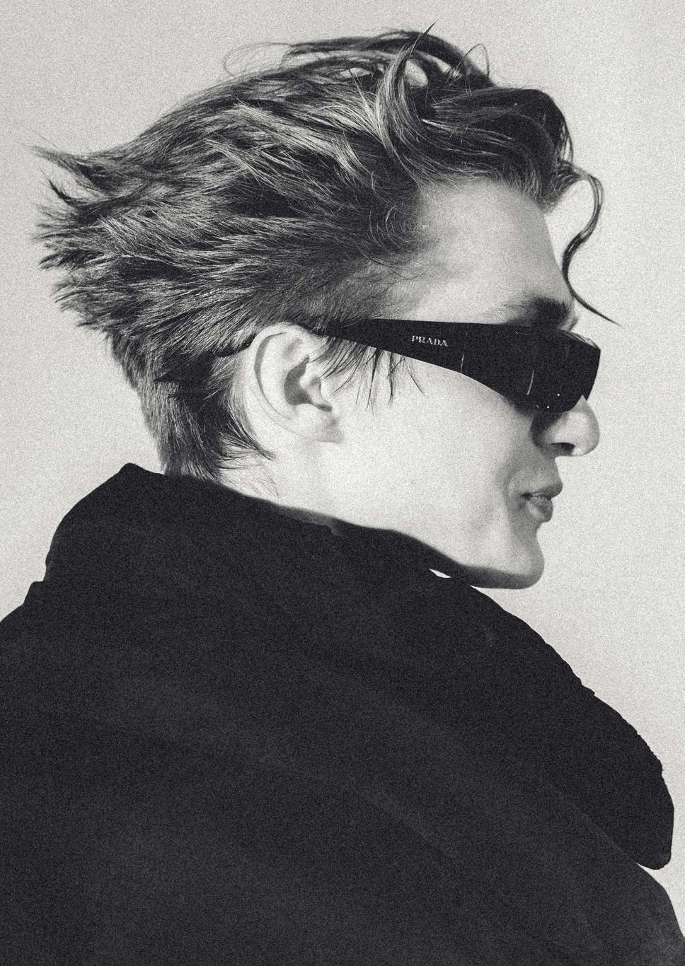Profil en noir et blanc d’un modèle aux cheveux mi-longs séchés vers l’arrière avec des mèches libres ; fines lunettes de soleil noires et manteau noir à grand col.